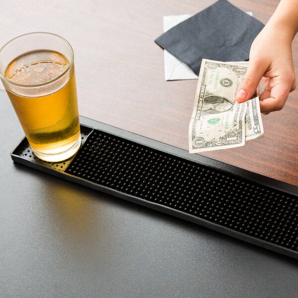 A person's hand holding a dollar bill over a black bar mat with small balls on it next to a glass of beer.