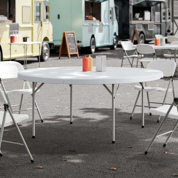 A round, white plastic bi-folding table with gray legs set up outdoors with folding chairs around it.