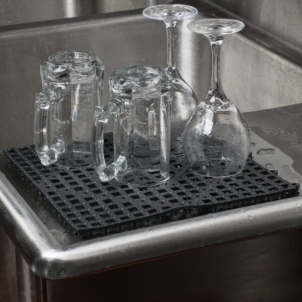 A group of glasses and cups on a black Choice interlocking bar mat on a counter.