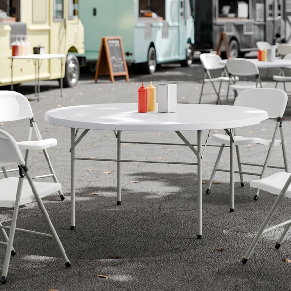 A round, white plastic bi-folding table with gray legs, set outdoors with folding chairs around it.