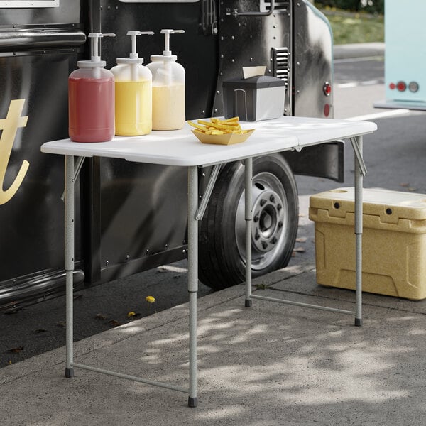 A 24-inch by 48-inch granite white plastic bi-folding table with gray adjustable height legs, set up outdoors near a food truck.