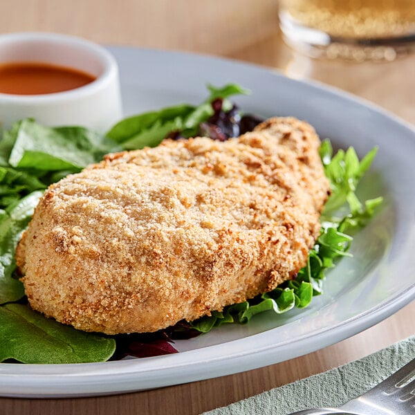 A white plate topped with a piece of breaded chicken on a table with a salad and a glass of liquid.