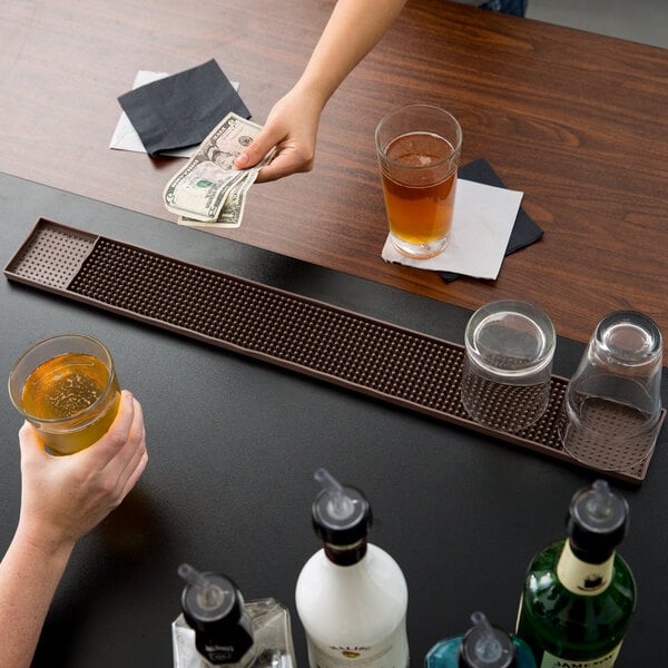 A person holding a glass of brown liquid and money on a brown bar mat.