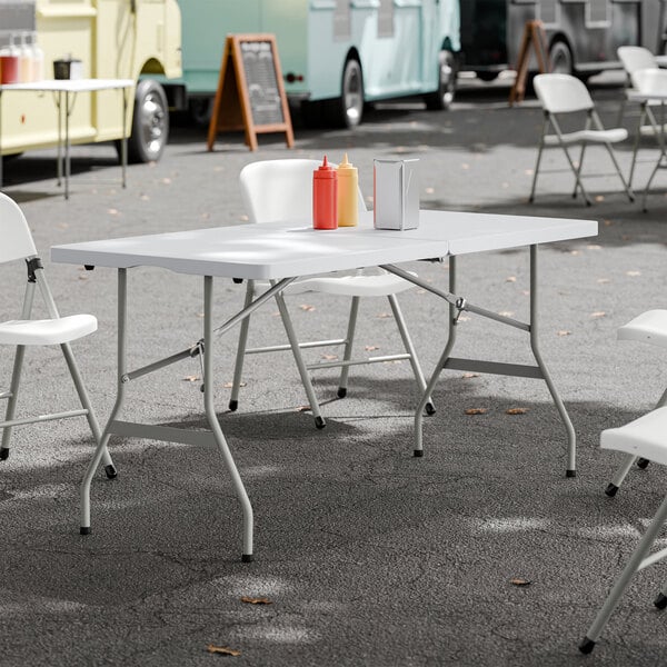 A 30 inch by 60 inch granite white plastic bi-folding table with gray legs, set up outdoors with condiment bottles and napkins on top.
