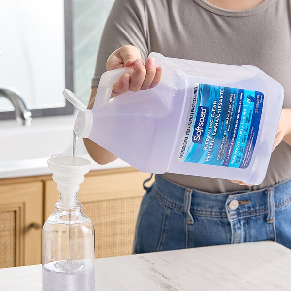 A woman pouring Softsoap liquid hand soap from a jug into a clear plastic bottle.