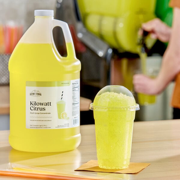 A gallon jug of Hometown Provisions Kilowatt Citrus Slushy concentrate next to a cup of yellow citrus slush.