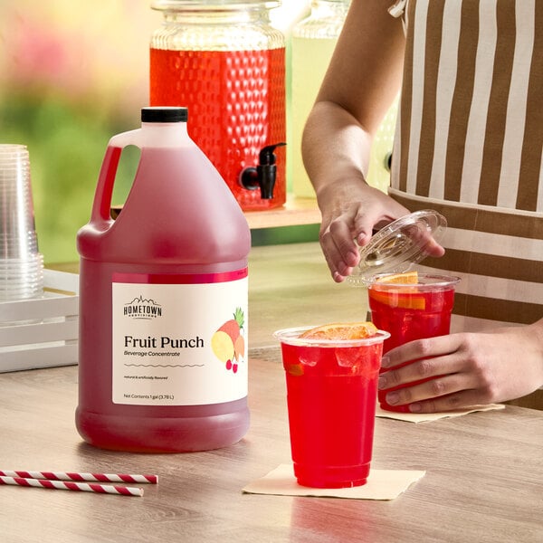 A gallon jug of Hometown Provisions Fruit Punch Beverage Concentrate placed on a table next to cups of prepared fruit punch.