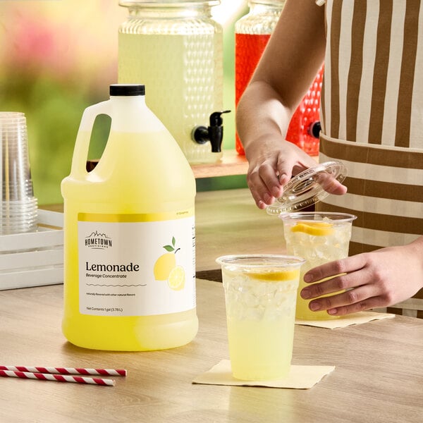 A gallon jug of Hometown Provisions Lemonade Beverage Concentrate on a counter next to cups of prepared lemonade with ice and lemon slices.