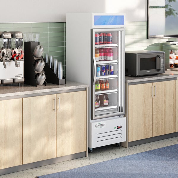 A white swing glass door merchandiser refrigerator with multiple shelves, stocked with beverages, situated between countertops in a break room or kitchen area.