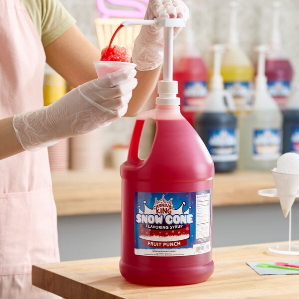 A gallon jug of Carnival King Fruit Punch Snow Cone Syrup with a pump dispenser being used to pour syrup onto a snow cone.