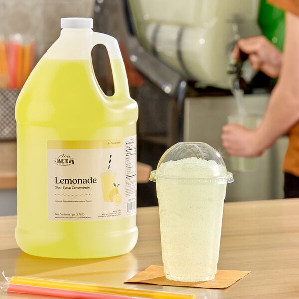A gallon jug of Hometown Provisions Lemonade Slushy 5:1 concentrate next to a cup of prepared lemonade slush.