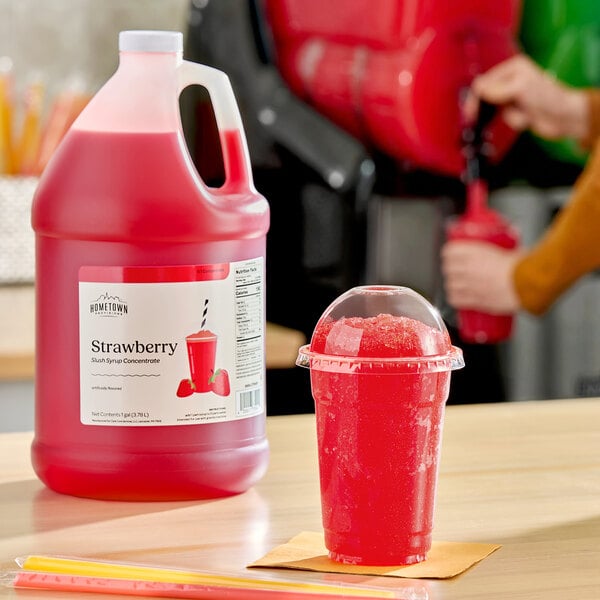 A gallon jug of Hometown Provisions strawberry slush syrup concentrate next to a cup of red strawberry slush.