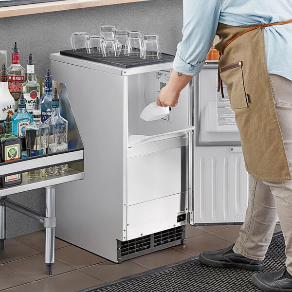A man in an apron standing next to a Hoshizaki undercounter ice machine on a counter with a clear glass of liquid.