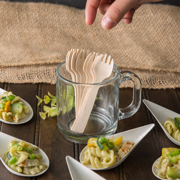 A glass mug filled with wooden taster forks resting on a plate of pasta and vegetables.