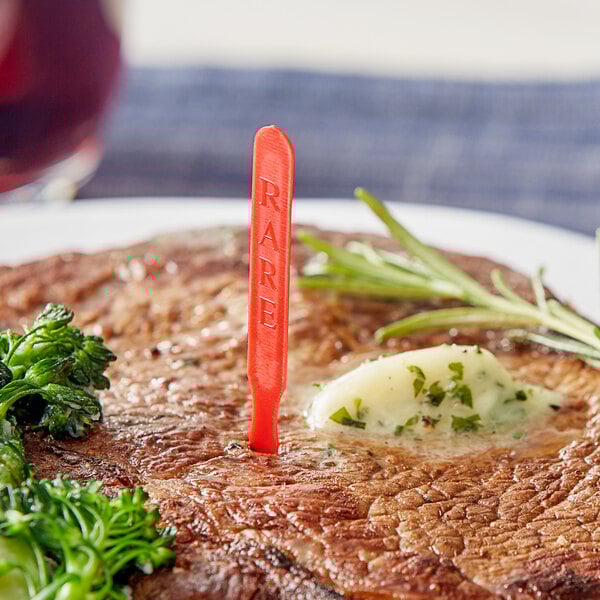 A steak with a Royal Paper red rare steak marker in the middle of it on a plate with broccoli and a fork.