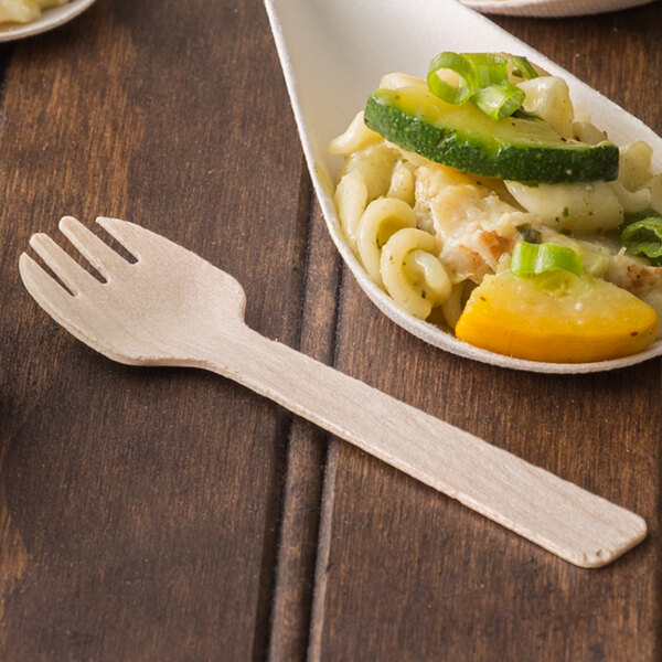 A wooden taster fork next to a plate of pasta with vegetables.