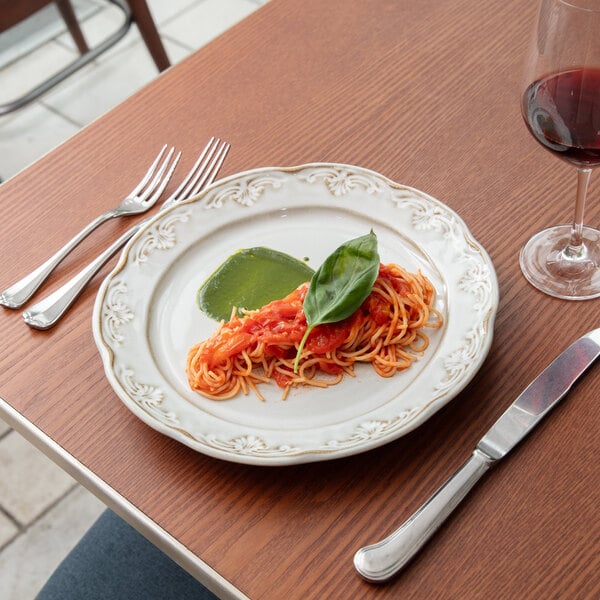 A white round stoneware charger plate with a scalloped edge and embossed pearl design, shown with a serving of pasta and a green sauce.