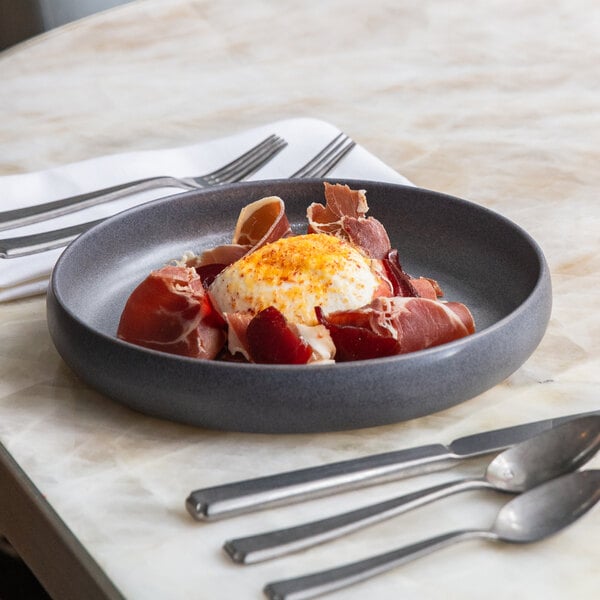 A round, volcanic gray stoneware serving plate with a raised rim, shown holding a dish of cured meat and a poached egg.
