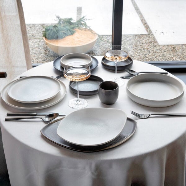 A set of white round raised rim coupe stoneware plates arranged on a table with other dinnerware and glassware.