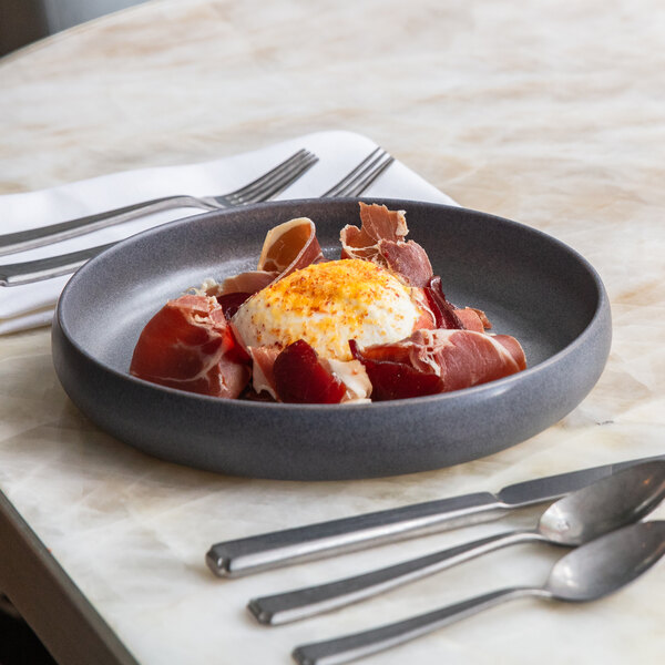 A round volcanic gray stoneware plate with a raised rim, holding a dish of cured meat and a poached egg, placed on a marble table with cutlery nearby.