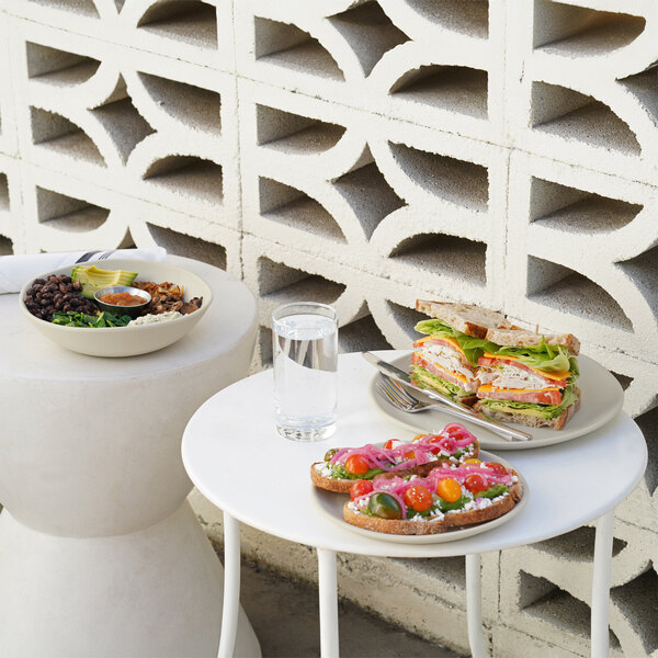 A round ivory melamine plate holding a sandwich, displayed on a white table next to a glass of water and another plate of food.