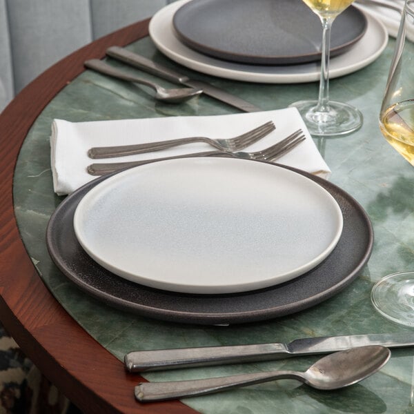 A round, thermal white stoneware salad plate placed on a dark charger at a set dining table.