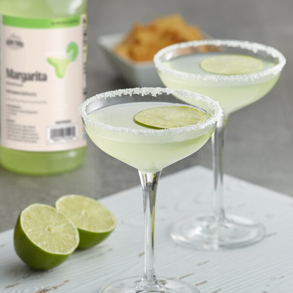 A bottle of Hometown Provisions Margarita Mix next to two margarita glasses with salted rims and lime slices.