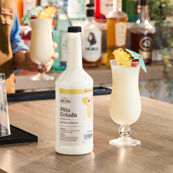 A 1-liter bottle of Hometown Provisions Piña Colada mix placed on a bar counter next to a prepared piña colada drink garnished with pineapple and a cocktail umbrella.