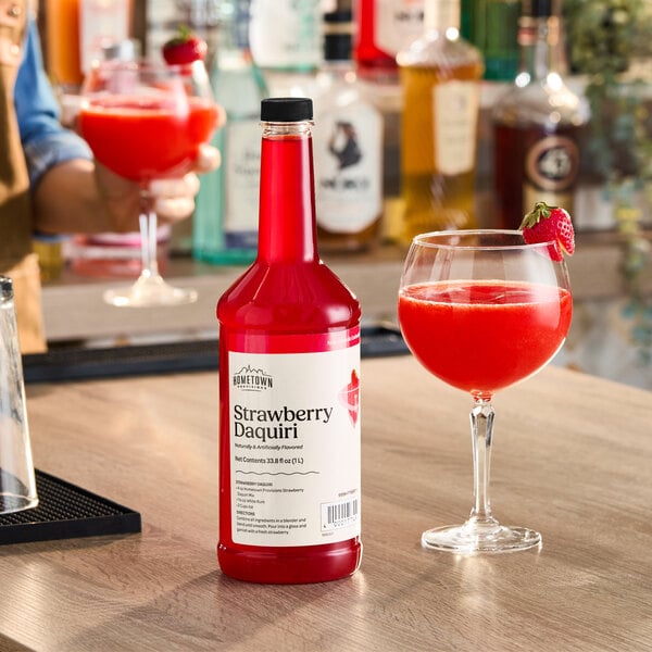 A bottle of Hometown Provisions Strawberry Daiquiri Mix next to a glass of strawberry daiquiri garnished with a strawberry.