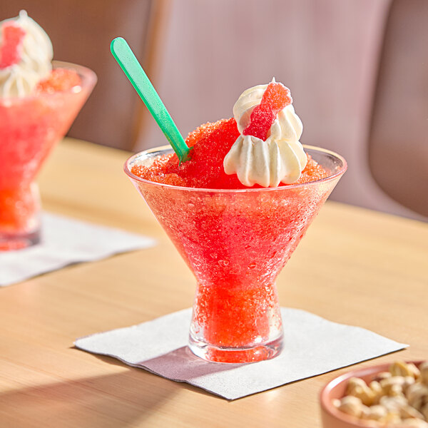 A red slushy served in a clear glass, topped with whipped cream and a gummy candy, with a green spoon.