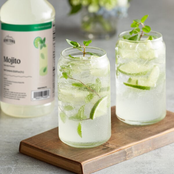 A bottle of Hometown Provisions Mojito Mix and two glasses of mojito garnished with lime and mint on a wooden board.
