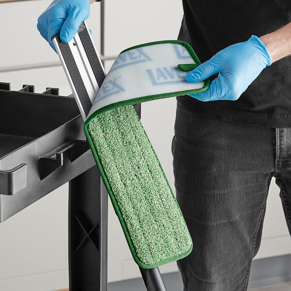 A person wearing blue gloves using a green and white Lavex microfiber mop pad to clean a table in a professional kitchen.