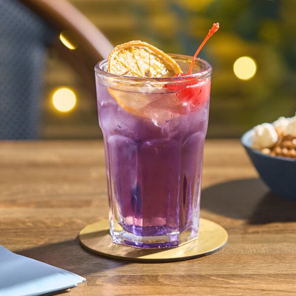 An Acopa Memphis highball glass of purple liquid with a cherry and orange slice on a table with a bowl of popcorn and a coaster.
