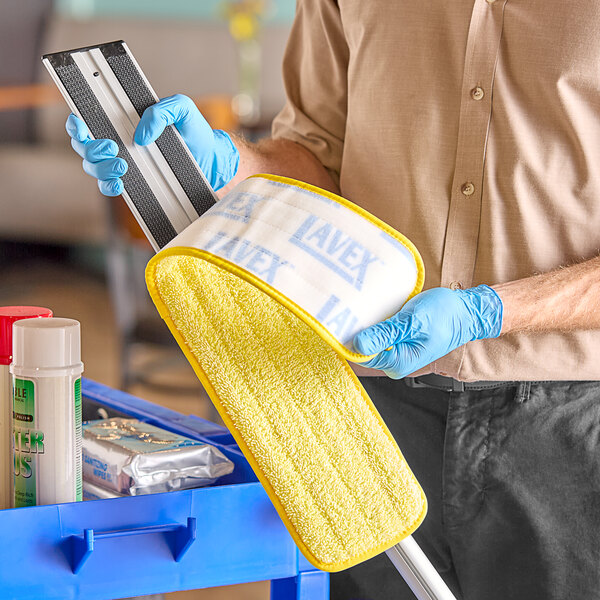 A person wearing blue gloves using a yellow Lavex microfiber mop pad to clean a floor.