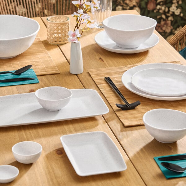 A small white round melamine bowl placed on a wooden dining table with other tableware.