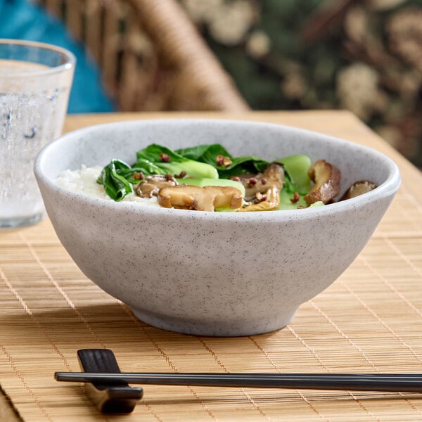 A light gray round melamine bowl filled with rice, vegetables, and mushrooms, placed on a bamboo mat with chopsticks and a glass of water nearby.