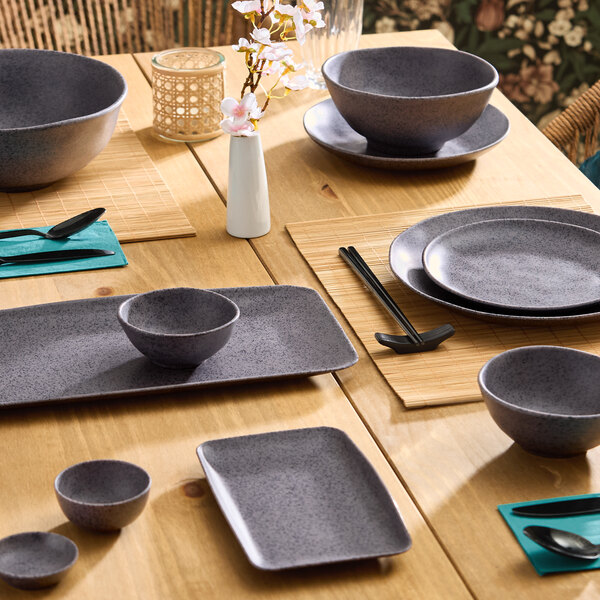 A set of small, dark gray round melamine bowls placed on a wooden dining table with matching dinnerware.