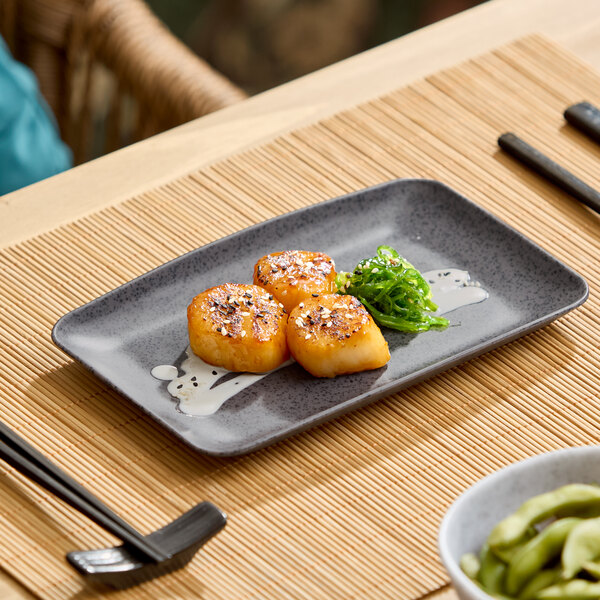 A dark gray rectangular melamine platter with three seared scallops and a small serving of seaweed salad.