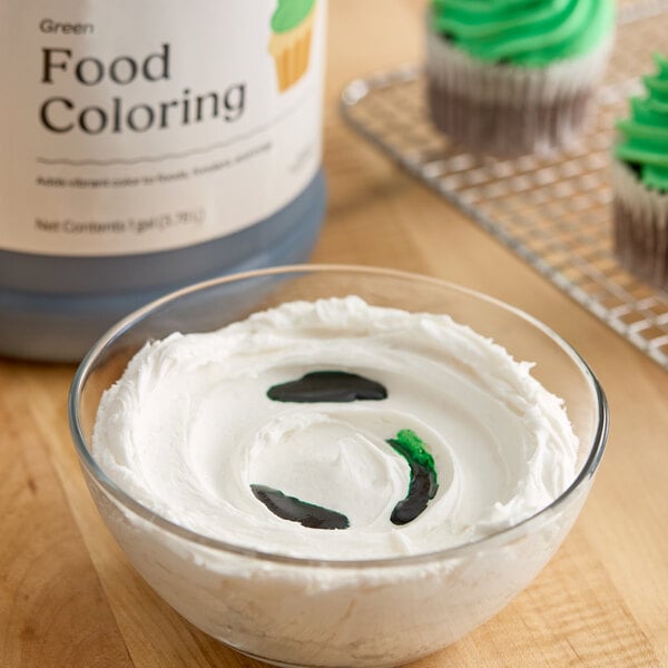 A bowl of white frosting with green food coloring being added, next to a large container of green food coloring and cupcakes with green frosting in the background.