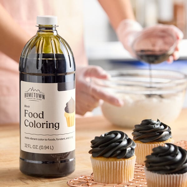 A 32 fl. oz. bottle of Hometown Provisions black food coloring placed on a kitchen counter near cupcakes with black frosting.