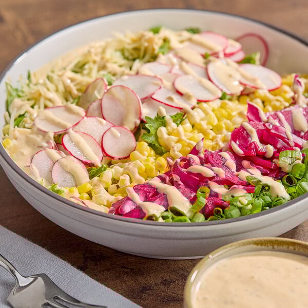 A bowl of salad topped with sliced radishes, corn, green onions, and a creamy dressing, with a small bowl of dressing on the side.