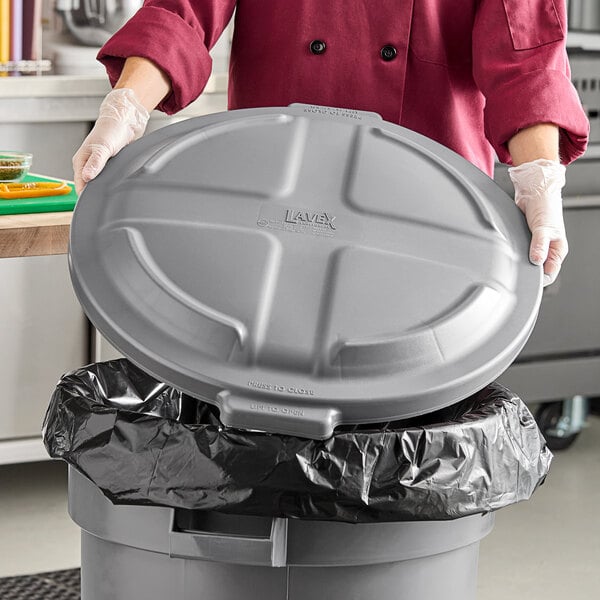 A gray round commercial trash can lid being placed on a trash can.