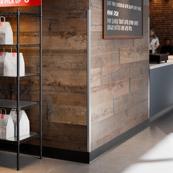 A stainless steel wall corner guard with mounting holes installed on the corner of a wooden wall in a commercial setting.