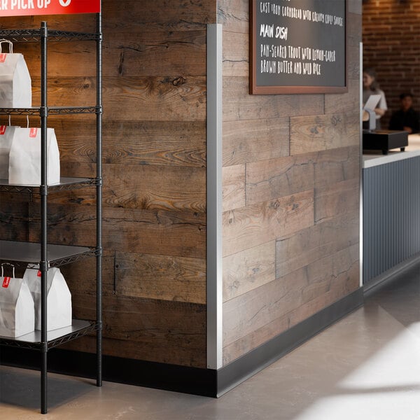 A stainless steel wall corner guard with mounting holes installed on the corner of a wooden wall in a commercial setting.