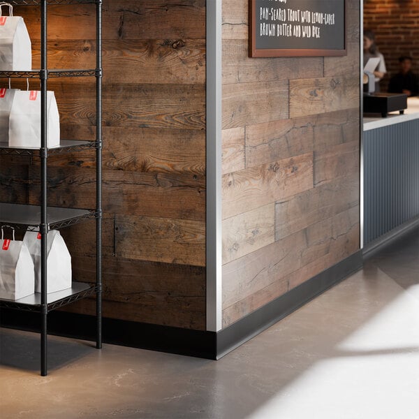 A stainless steel wall corner guard with adhesive strips installed on the corner of a wooden wall in a commercial setting.