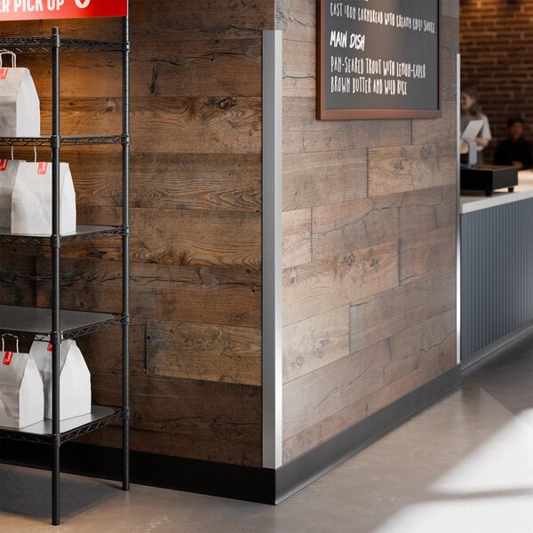 A stainless steel wall corner guard with adhesive strips installed on a wooden wall corner in a commercial setting.