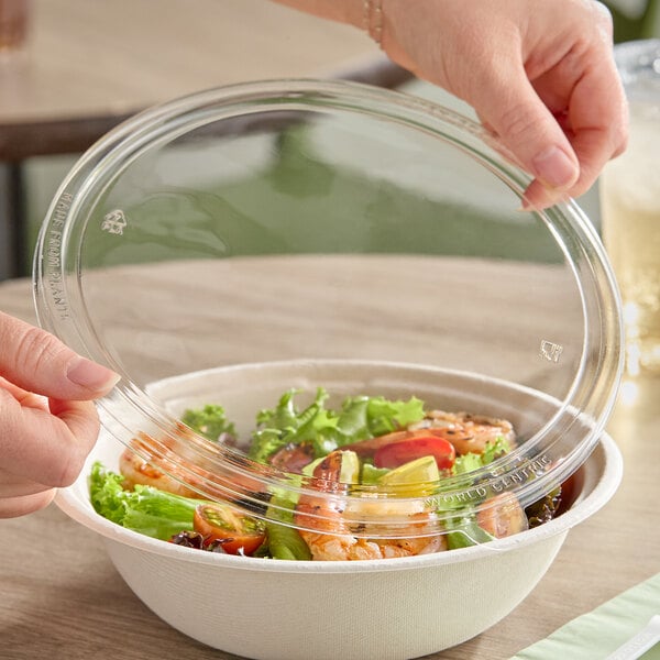 A clear compostable plastic lid being placed on a wide fiber take-out bowl filled with salad.