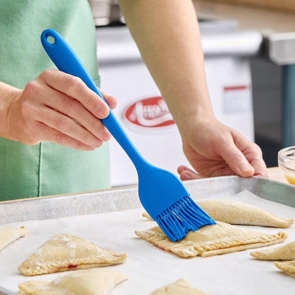A blue silicone basting or pastry brush being used to apply a coating to pastries on a baking sheet.