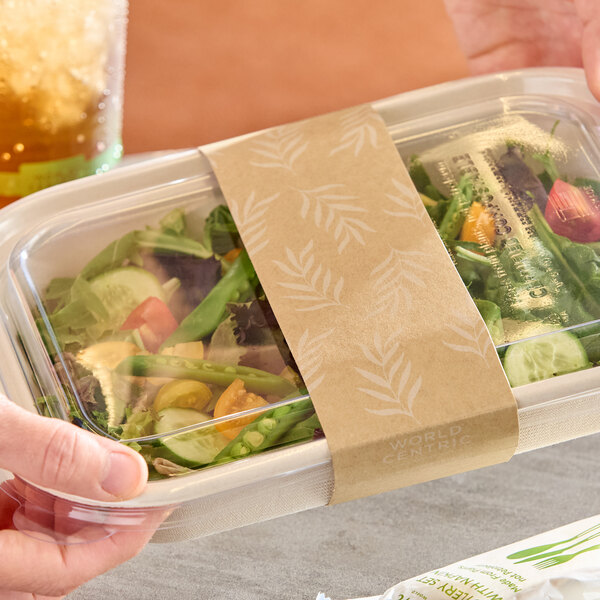 A compostable paper sleeve wrapped around a clear-lidded fiber food container filled with salad.