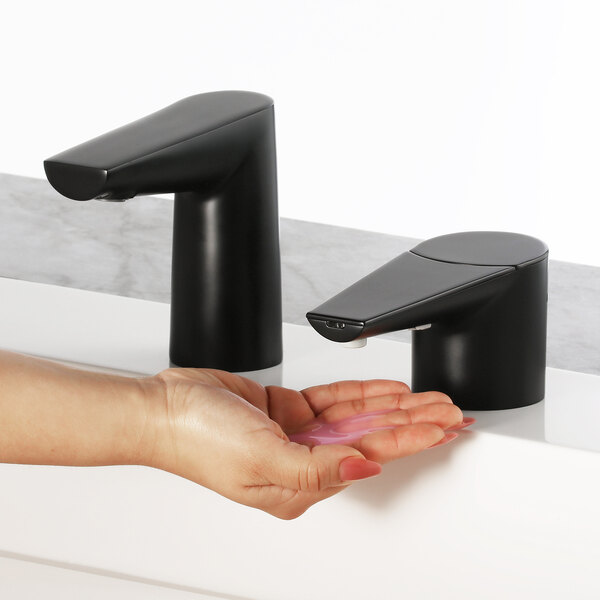 A person's hand pressing a Bobrick matte black deck-mount automatic liquid soap dispenser over a sink.